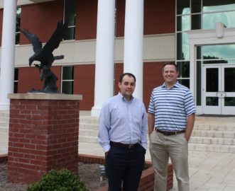 l-r William Cooper and Justin Boyd pose in front of the Harris College of Business building next to a bronzed eagle. They are pursuing their PhDs.