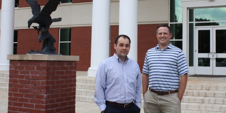 l-r William Cooper and Justin Boyd pose in front of the Harris College of Business building next to a bronzed eagle. They are pursuing their PhDs.