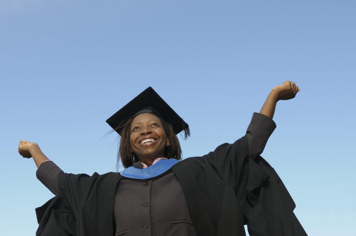 New grad wearing cap and gown triumphantly raises arms 