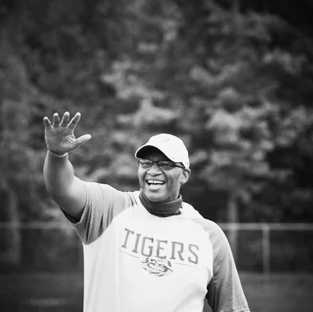 Dr. Gregory Moore raises his hand during an athletic event. He's wearing a Tigers shirt and a baseball cap.