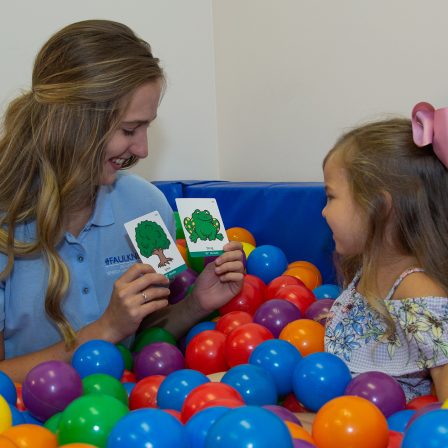 Speech-language pathologist sitting in ball pit with child and holding up pictures of tree and frog