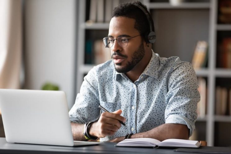 Student wearing headphones studying on laptop while taking handwritten notes 