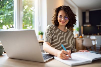 Young woman sitting in front of computer and writing in notebook