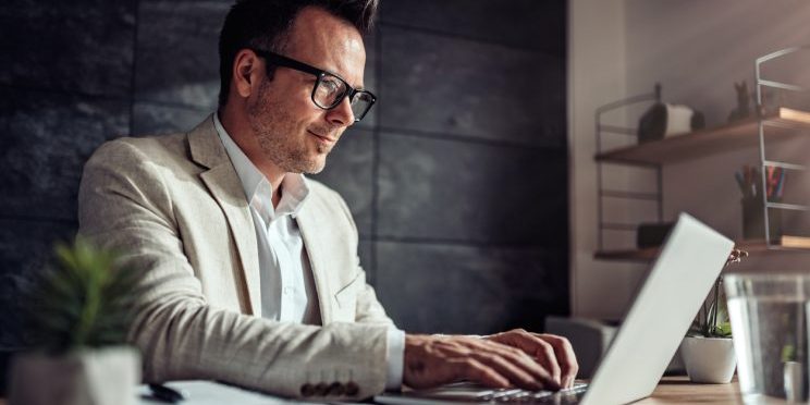 Businessman sitting at his desk and using laptop in the office Graduate student in blazer and glasses working on laptop at desk