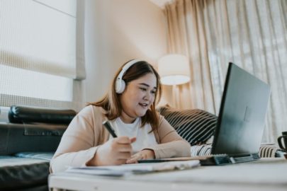 Student wearing headphones looking at laptop screen and taking handwritten notes